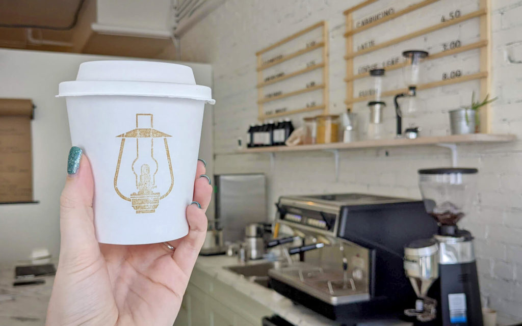Hand Holding White To Go Cup with Lightbulb Logo in Front of Coffee Equipment