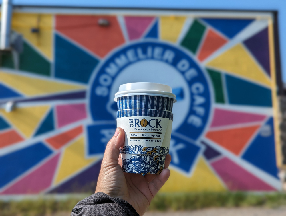 Hand raising an Old Rock Roastery cup of coffee in Sudbury in front of a colourful mural